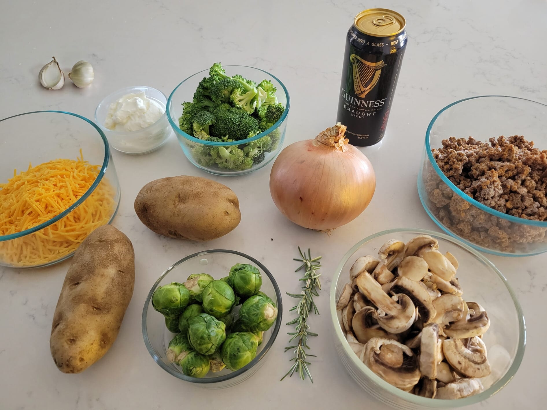 chopped broccoli, brussels sprouts, soy crumbles, sliced mushrooms, Greek yogurt, and shredded cheddar cheese in clear bowls on top of a white marbled countertop. There are also one whole yellow onion, two whole russet potatoes, a sprig of rosemary, and a black can of Guinnes Draught Stout